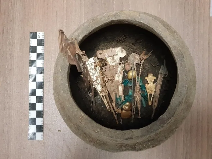 Gold figurines, beads and green stones inside a Muisca offering vessel, photographed with a scale bar